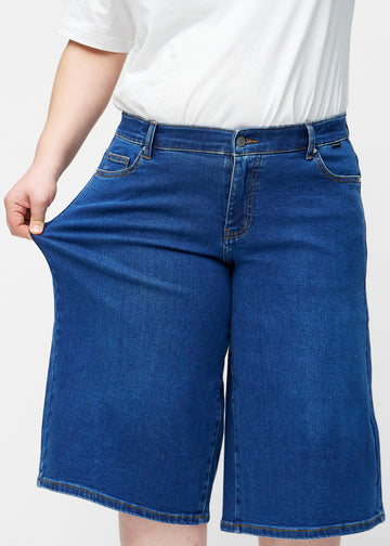 Plus-size model strækker de løse, mørkeblå denim capris i farven Oceans ved hoften for at vise materialets stretch.