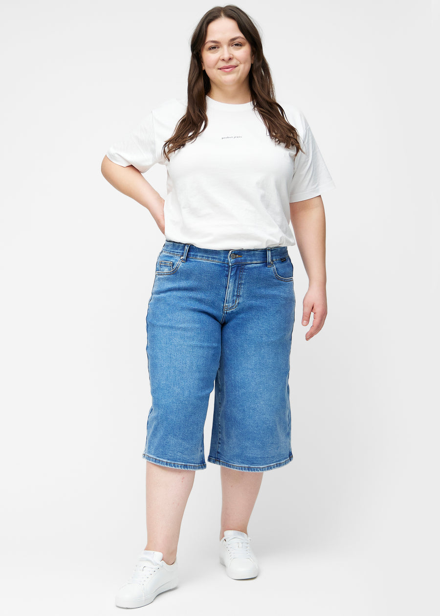 Fuldt billede af plus-size model i løse, klassisk blå denim capris i farven Rivers, der går til lige under knæet.