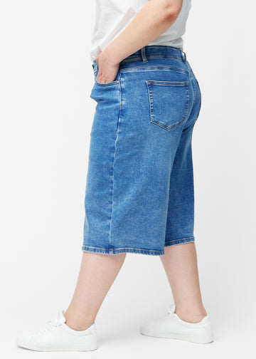 Løse, klassisk blå denim capris i farven Rivers set fra siden på plus-size model, hvor den brede pasform og længden til lige under knæet er synlig.