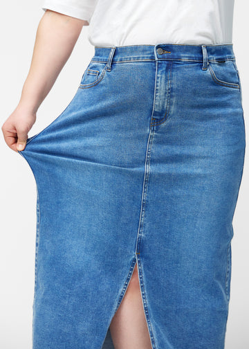 Plus-size model strækker den lange, klassisk blå denimnederdel i farven Rivers ved hoften for at vise materialets stretch.