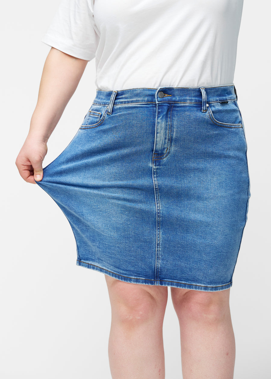Plus-size model strækker klassisk blå, tætsiddende og knælang denimnederdel ved låret for at vise stretch.