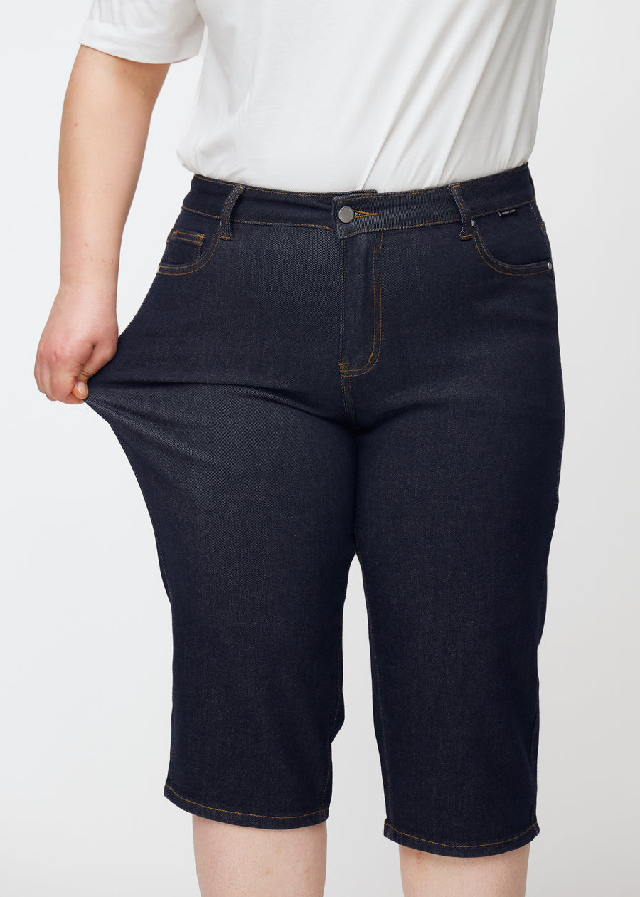 Plus-size model stretches deep dark blue denim capris for women in the colour Midnights at the thigh to show stretch.