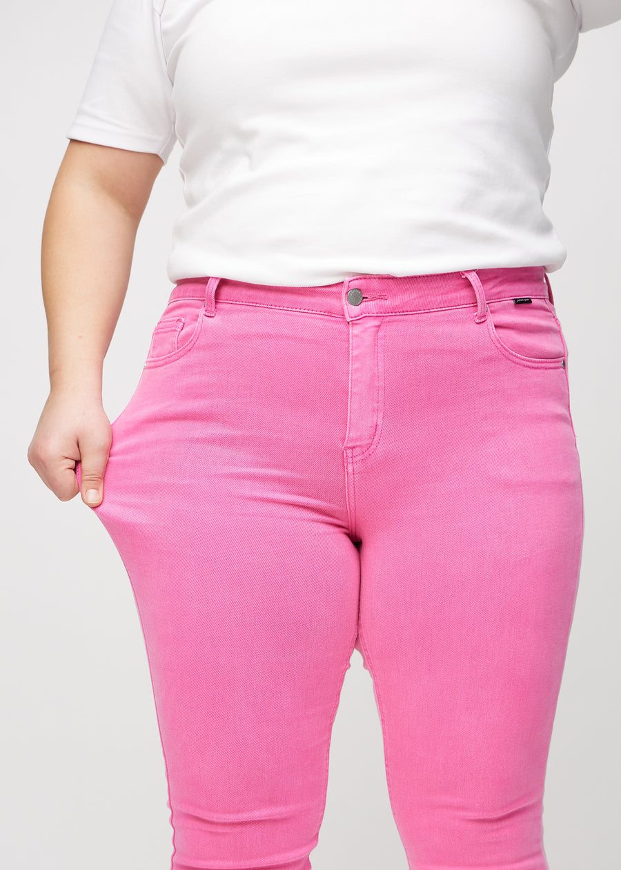 Skinny jeans for women in light pink, color Flamingos. A plus-size model stretches the jeans at the thigh to show the fabric&