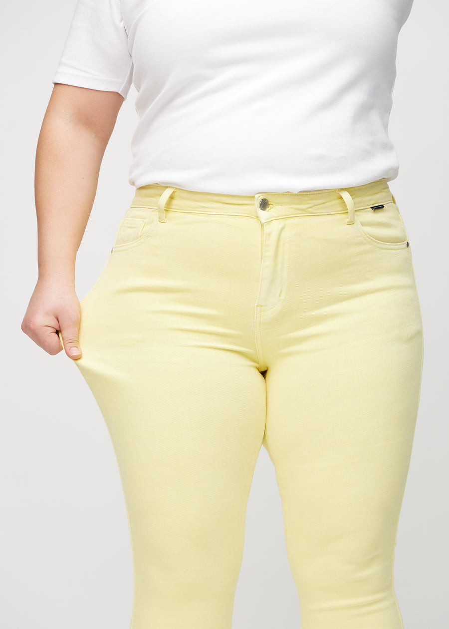 Skinny jeans for women in light yellow, color Lemonades. A plus-size model stretches the jeans at the thigh to show the fabric&