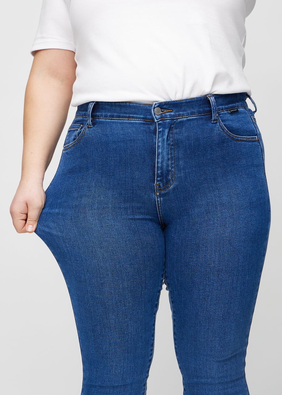 Skinny jeans for women in dark blue, color Oceans. A plus-size model stretches the jeans at the thigh to show the fabric&