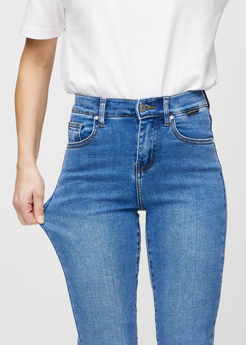 Slim jeans for women in blue, color Rivers. A model stretches the jeans at the thigh to show the fabric&