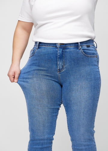 Slim jeans for women in blue, color Rivers. A plus-size model stretches the jeans at the thigh to show the fabric&