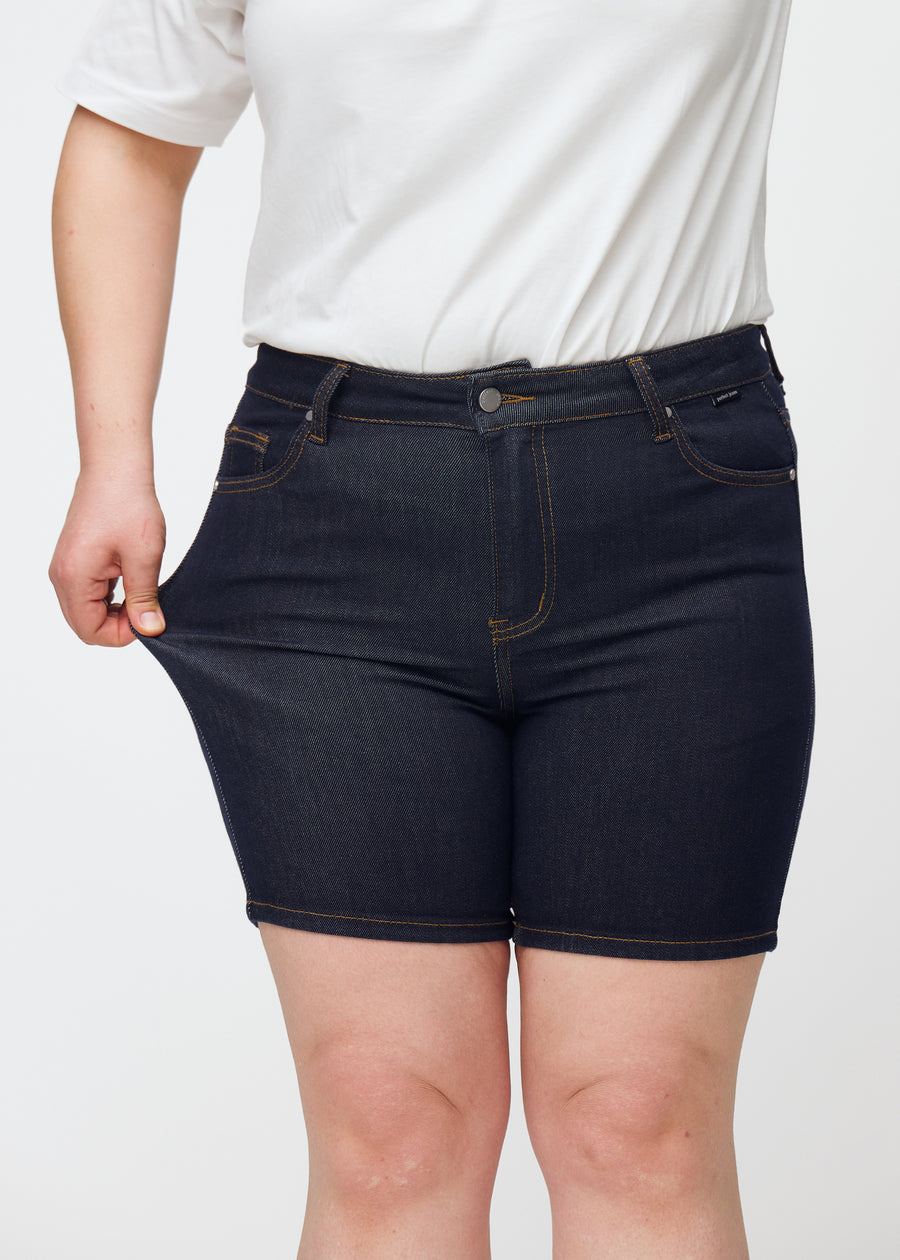 Plus-size model stretches deep dark blue denim shorts for women in the colour Midnights at the thigh to show stretch.
